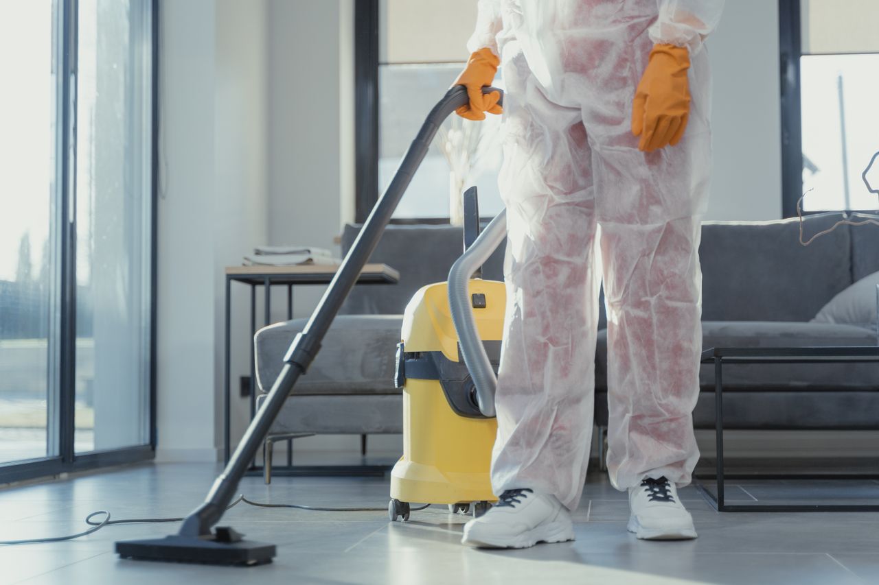 A cleaner wearing protective coveralls and gloves is vacuuming a contemporary living room.